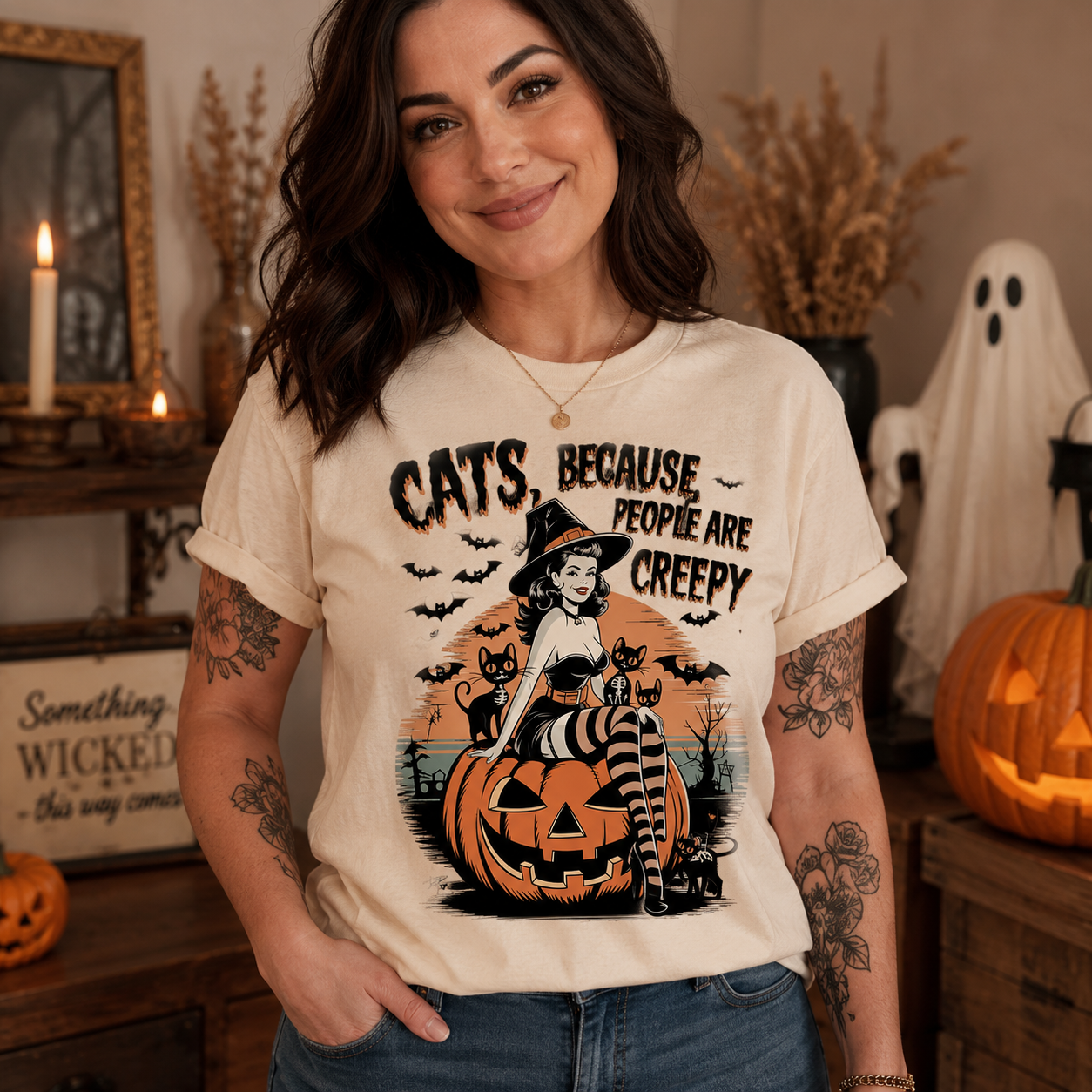 A woman wearing a cream-colored graphic t-shirt with a Halloween design featuring a witch, cats, and a pumpkin, posed in a cozy, decorated Halloween-themed room.