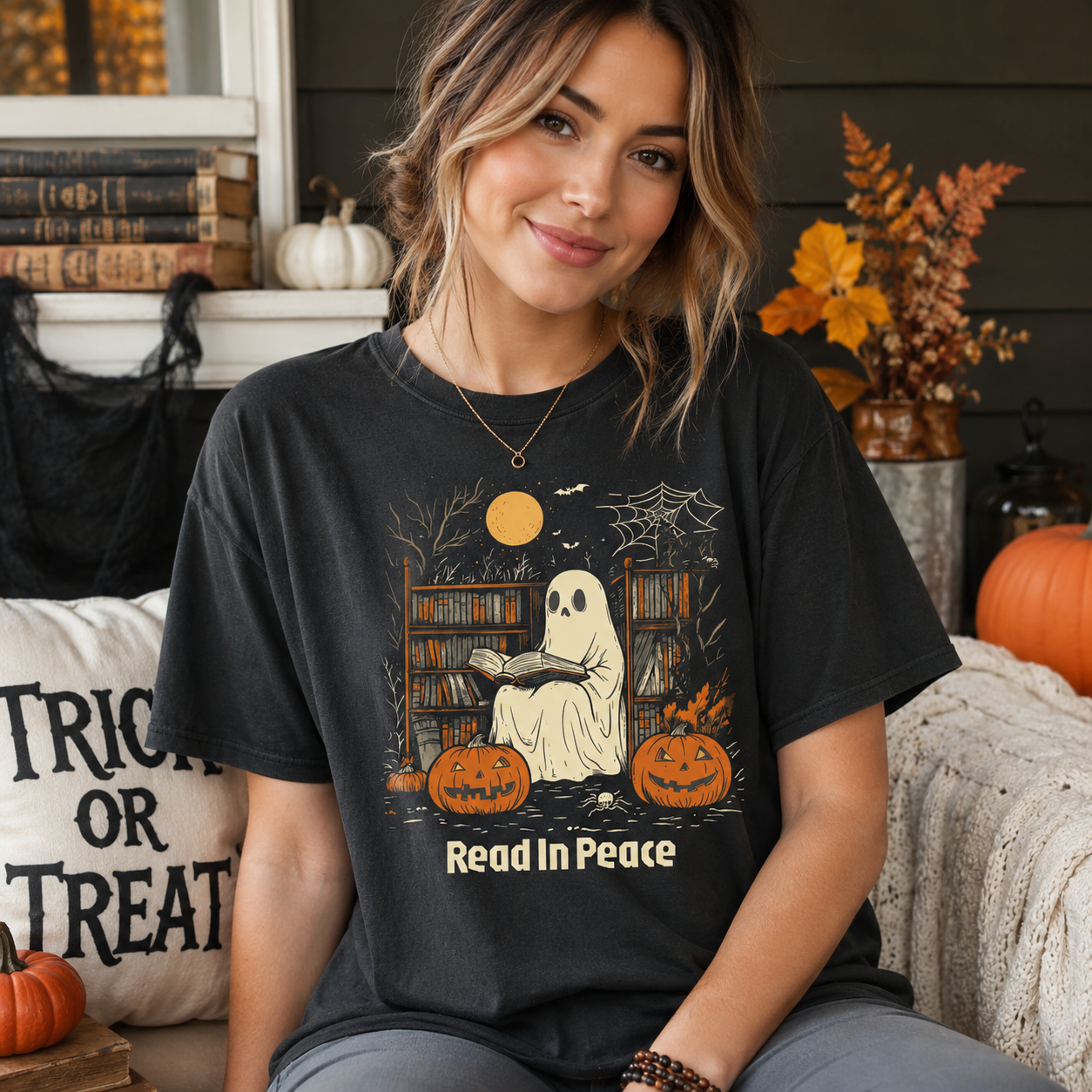 A woman wearing a black graphic t-shirt with a Halloween-themed ghost reading illustration and the text 'Read In Peace'. The background shows a cozy, decorated autumn setting with pumpkins, a 'Trick or Treat' pillow, and fall foliage.