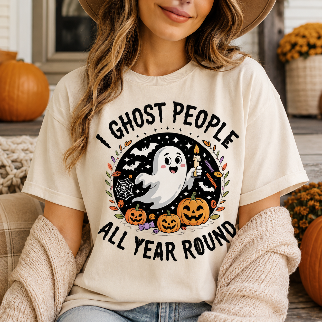 A woman wearing a cream-colored graphic t-shirt with a Halloween design that reads 'I GHOST PEOPLE ALL YEAR ROUND'. The graphic features a cartoon ghost, pumpkins, and spiderwebs.