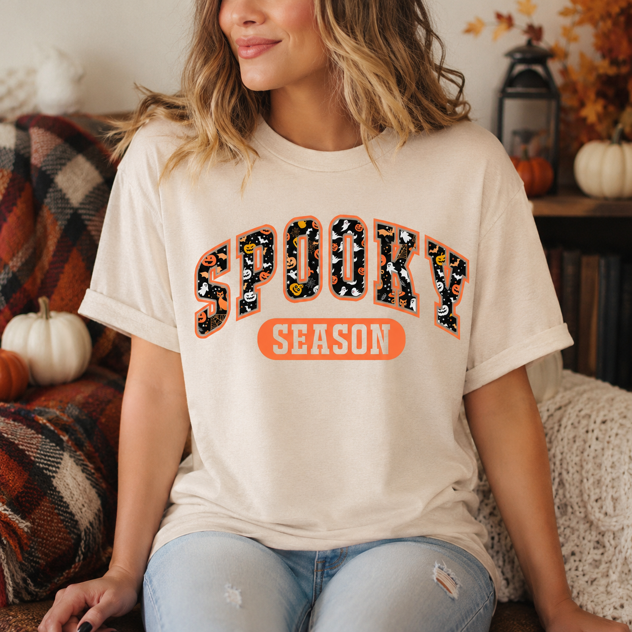 A woman wearing a cream-colored graphic t-shirt with 'SPOOKY SEASON' printed in a Halloween-themed patterned font, sitting in a cozy autumn-themed room.