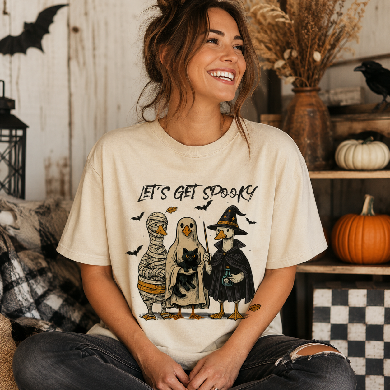 A woman wearing a cream-colored graphic t-shirt with a 'Let's Get Spooky' cartoon design of a mummy, a ghost, and a witch duck, posed in a cozy, rustic Halloween-themed room with pumpkins and a lantern.