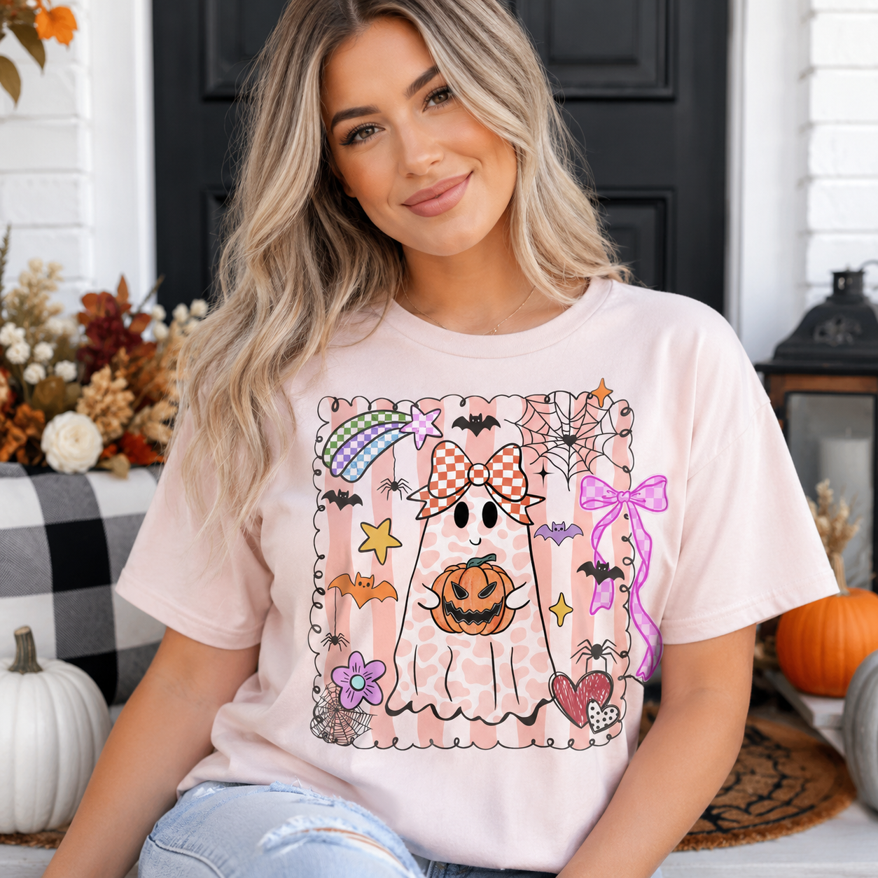 A woman wearing a light pink graphic t-shirt with a cute Halloween ghost design, posed in a cozy fall-themed setting with pumpkins and autumn decor.