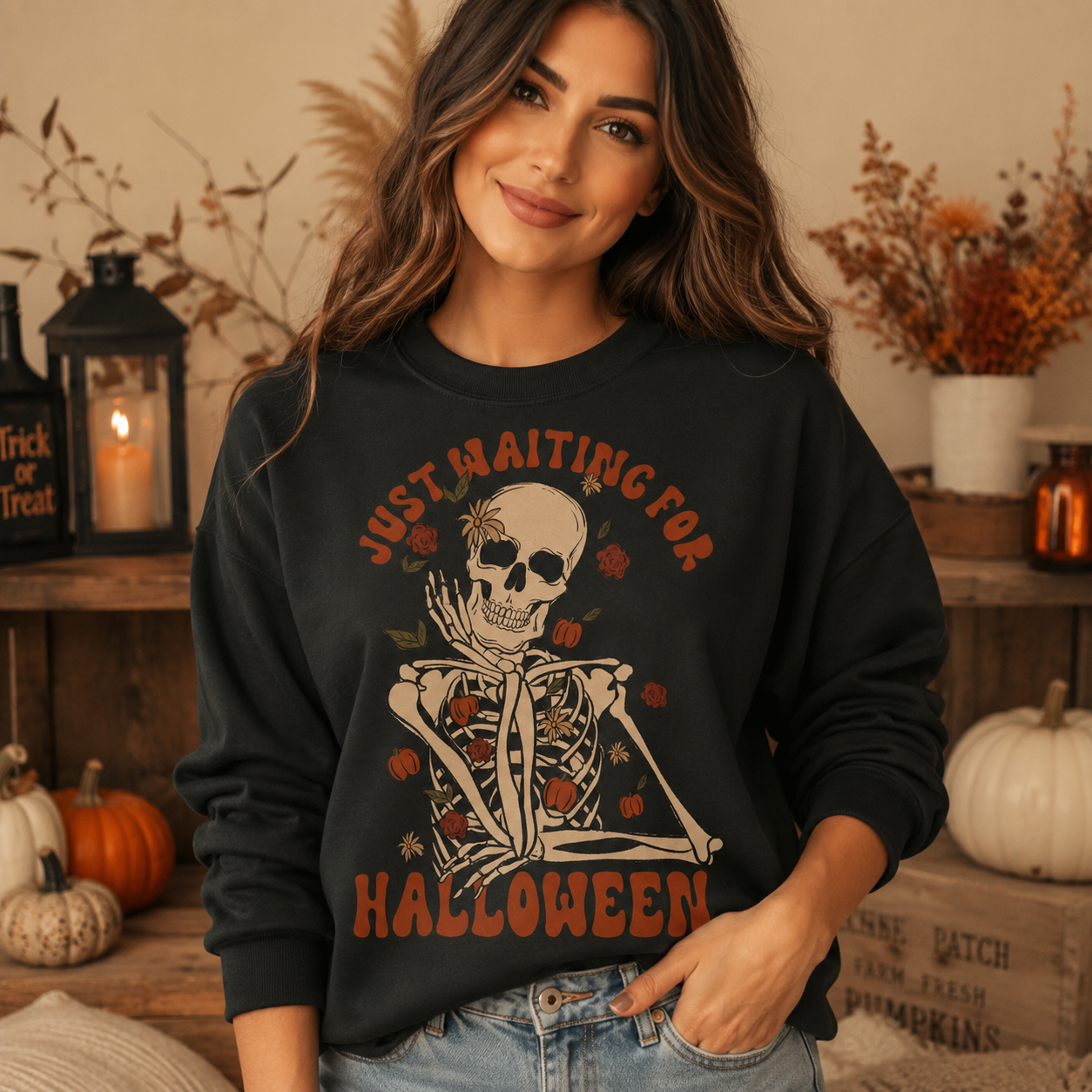 A woman wearing a black crewneck sweatshirt with a large skeleton graphic and the text 'JUST WAITING FOR HALLOWEEN' in orange, posed in a cozy, decorated autumn-themed room.