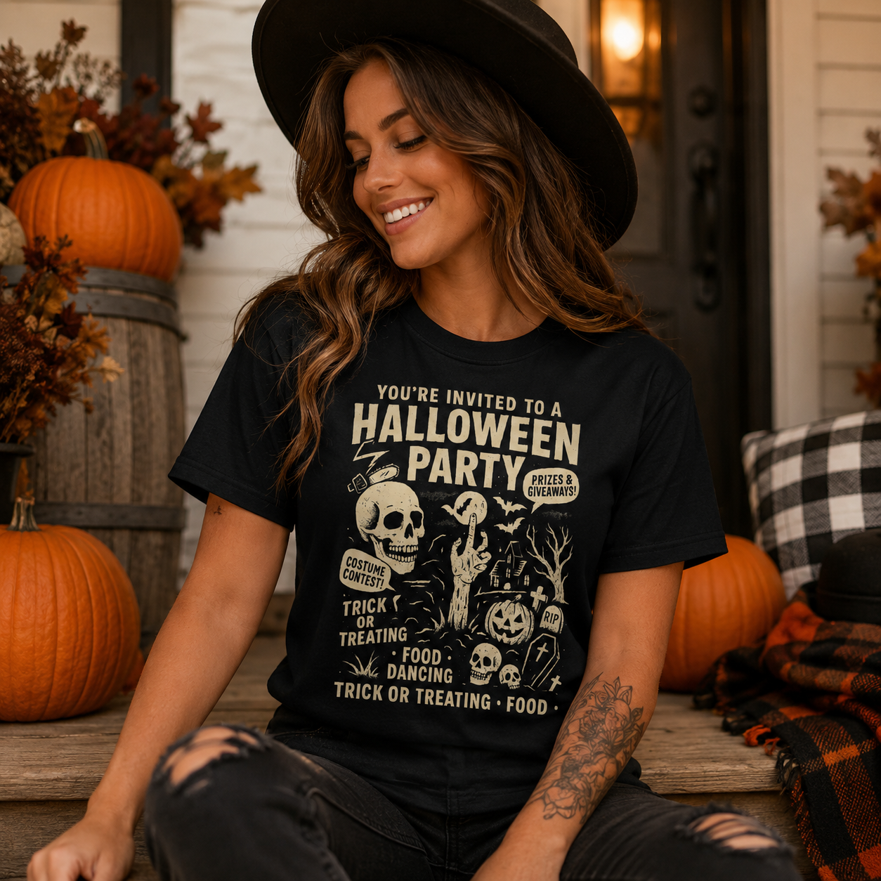 A woman wearing a black graphic t-shirt with a Halloween-themed design, sitting on a porch with pumpkins and autumn decorations in the background.