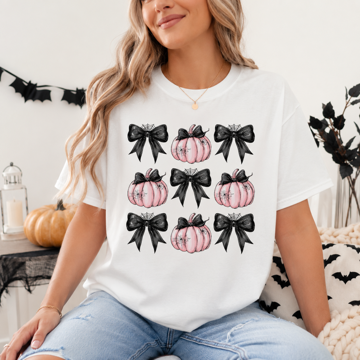 A woman wearing a white graphic t-shirt with a pattern of black bows and pink pumpkins, paired with light blue jeans, sitting in a cozy indoor setting with Halloween decorations.