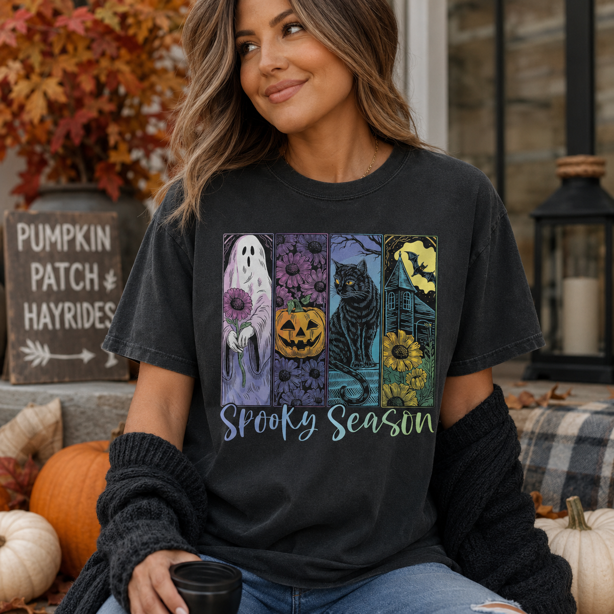 A woman wearing a dark charcoal gray graphic t-shirt with a 'Spooky Season' Halloween print, sitting in a decorated outdoor porch setting with pumpkins and autumn foliage.