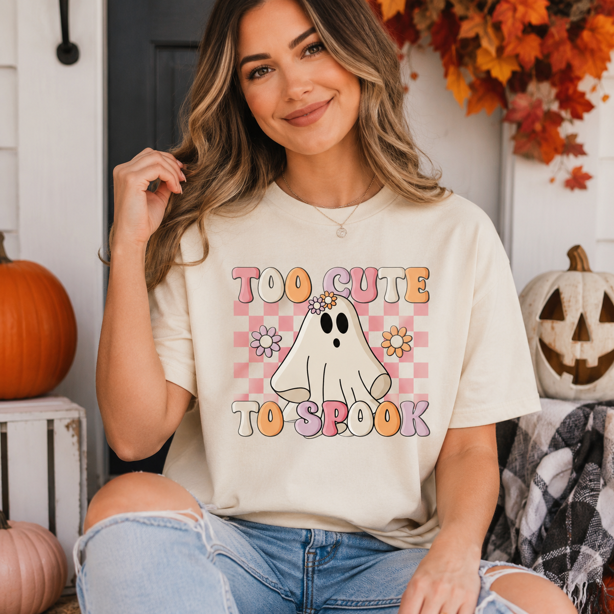 A woman wearing a cream-colored graphic t-shirt with a 'Too Cute to Suck' ghost design, paired with distressed blue jeans, posed in a cozy autumn-themed setting with pumpkins and fall leaves.