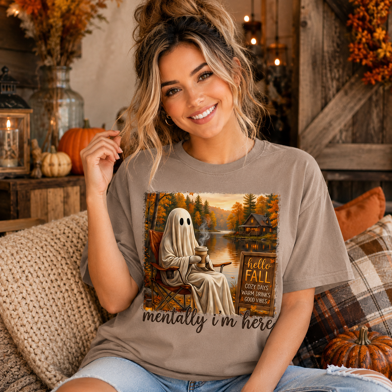 A woman wearing a taupe graphic t-shirt with a Halloween-themed ghost sitting by a lake and the text 'mentally i'm here', posed in a cozy autumn-themed room with pumpkins and a rustic wooden background.