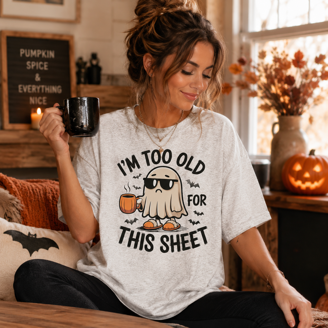 A woman sitting in a cozy, decorated room wearing a light gray graphic t-shirt with a cartoon ghost wearing sunglasses and the text 'I'M TOO OLD FOR THIS SHEET'. She is holding a black mug.