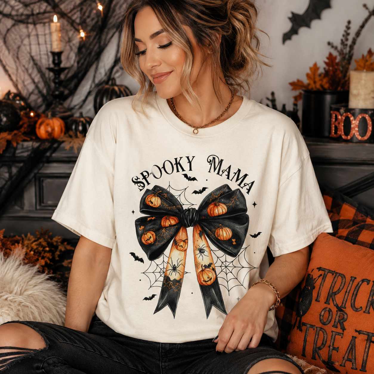 A woman wearing a cream-colored graphic t-shirt with a 'Spooky Mama' Halloween bow design, posed in a cozy, decorated autumn-themed room.