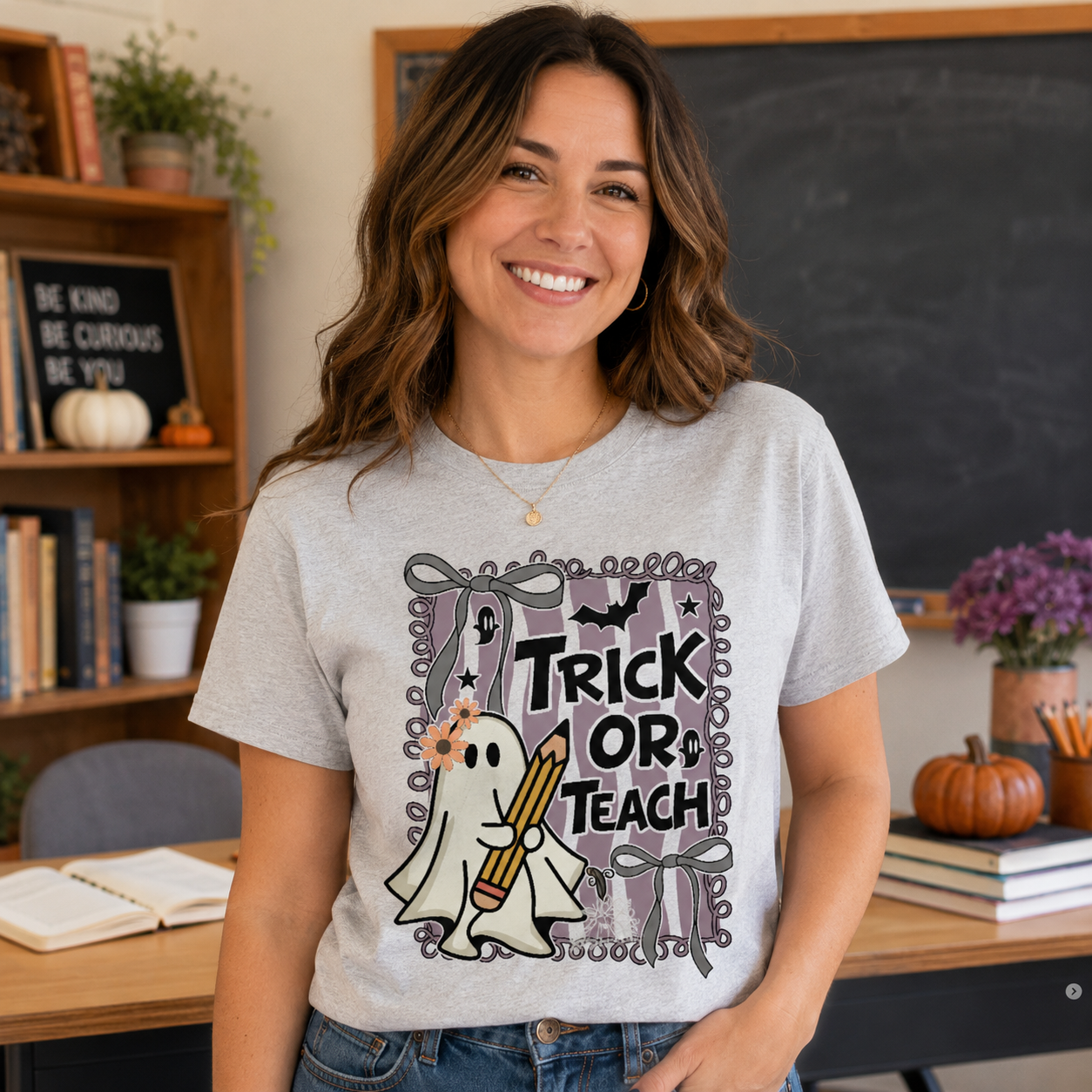 A smiling woman wearing a light gray graphic T-shirt with a 'Trick or Treat' ghost design, standing in a classroom setting with a chalkboard and shelves in the background.
