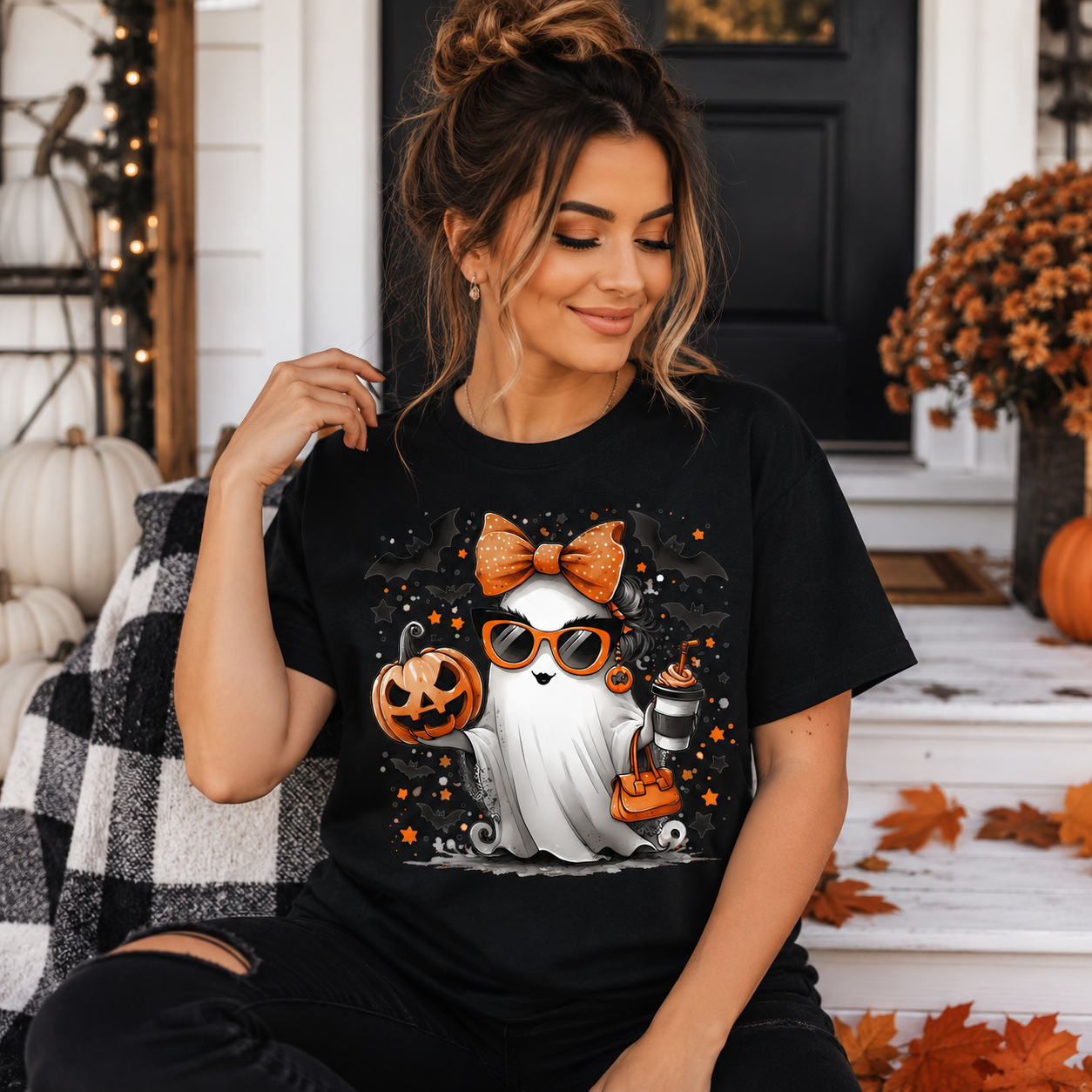 A woman wearing a black graphic t-shirt with a cute cartoon ghost wearing orange sunglasses and an orange bow, sitting on a porch decorated for Halloween.