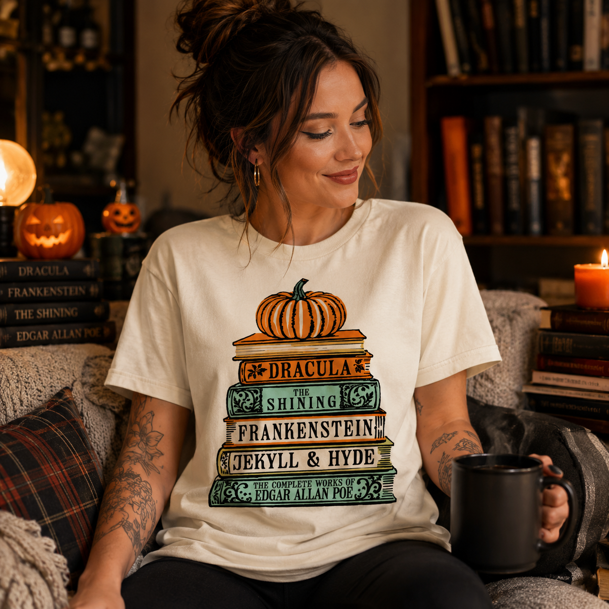 A woman sitting in a cozy, book-filled room wearing a cream-colored graphic t-shirt. The t-shirt features a colorful illustration of stacked books with pumpkins on top, labeled with Dracula, The Shining, Frankenstein, Jekyll & Hyde, and The Complete Works of Edgar Allan Poe.