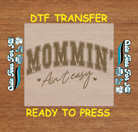 Retro varsity-style text reading “Mommin’ Ain’t Easy” in brown letters with hearts.