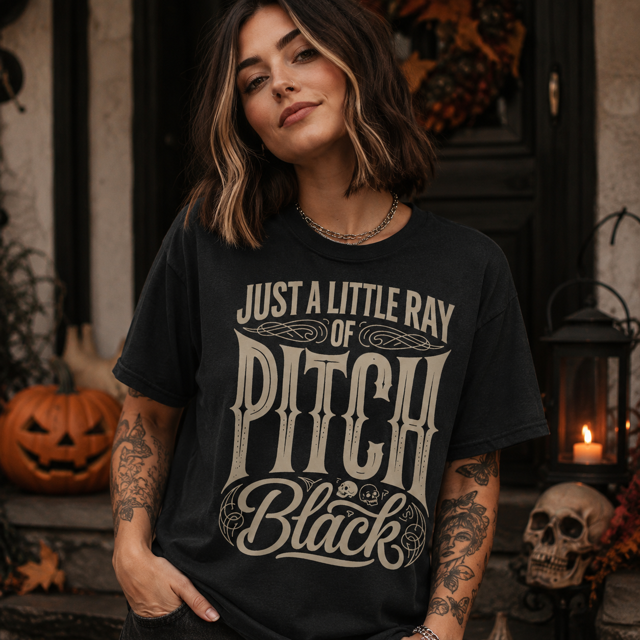 A woman wearing a black graphic t-shirt with the text 'JUST A LITTLE RAY OF PITCH BLACK' in a distressed cream font, posed in a spooky Halloween-themed setting with pumpkins, a skull, and a lantern.