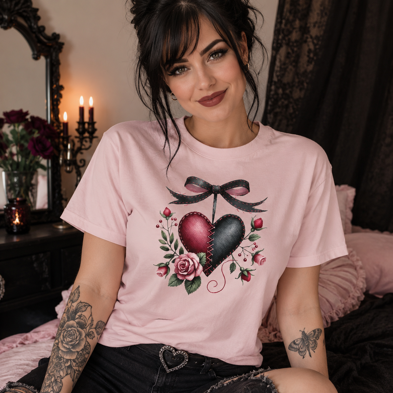 A woman wearing a light pink graphic t-shirt with a heart design, sitting in a bedroom setting with candles and a mirror in the background.