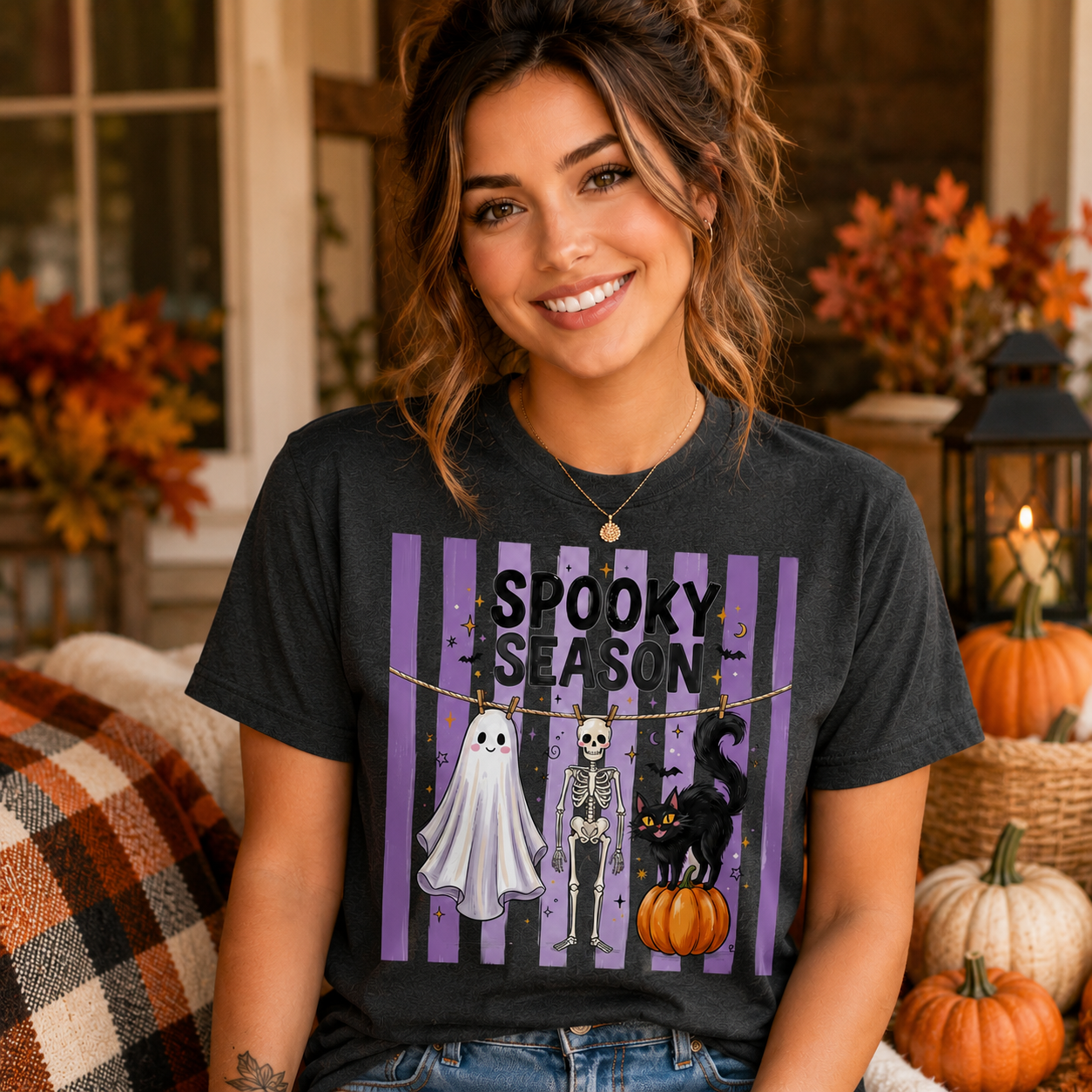 A woman wearing a dark charcoal gray graphic t-shirt with a purple and black 'SPOOKY SEASON' design featuring a ghost, skeleton, and black cat. She is posed in a cozy, autumn-themed indoor setting with pumpkins and fall foliage.