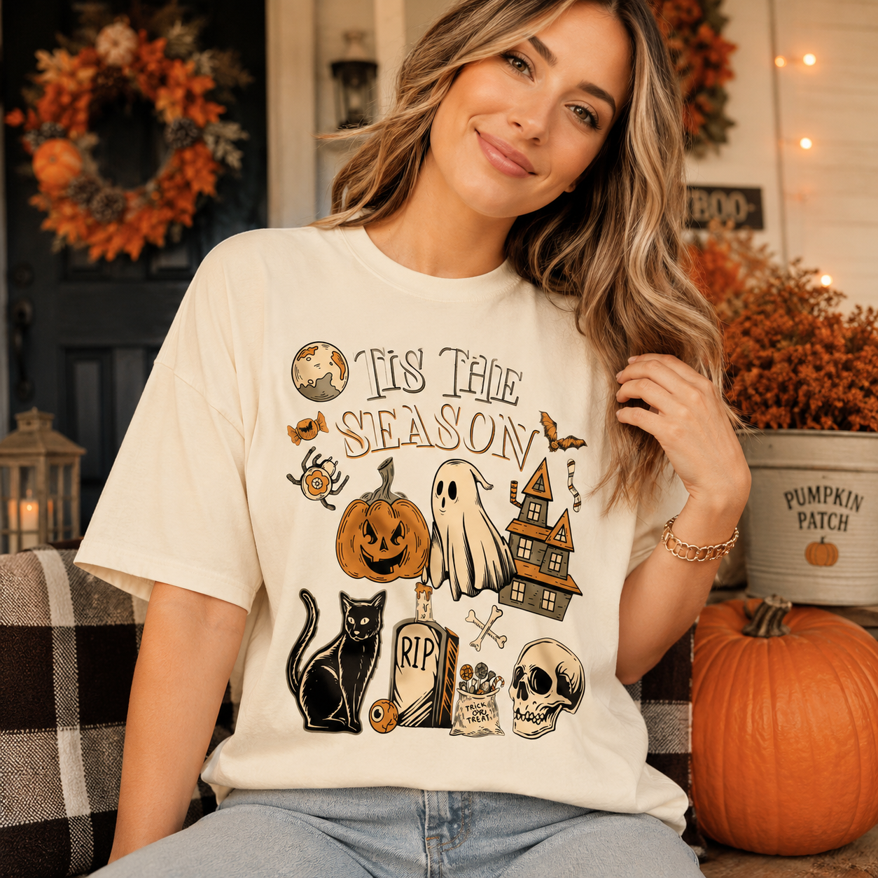 A woman wearing a cream-colored graphic t-shirt with a Halloween-themed print that reads 'Tis The Season'. She is sitting in a cozy, decorated home setting with pumpkins, a wreath, and fall decorations.