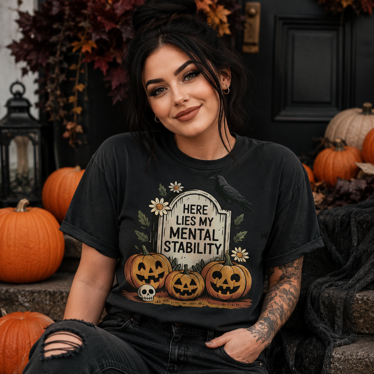 A woman wearing a black graphic t-shirt with a Halloween design that reads 'HERE LIES MY MENTAL STABILITY' above jack-o'-lanterns, a skull, and a crow. She is sitting in a decorated outdoor setting with pumpkins and a black door behind her.