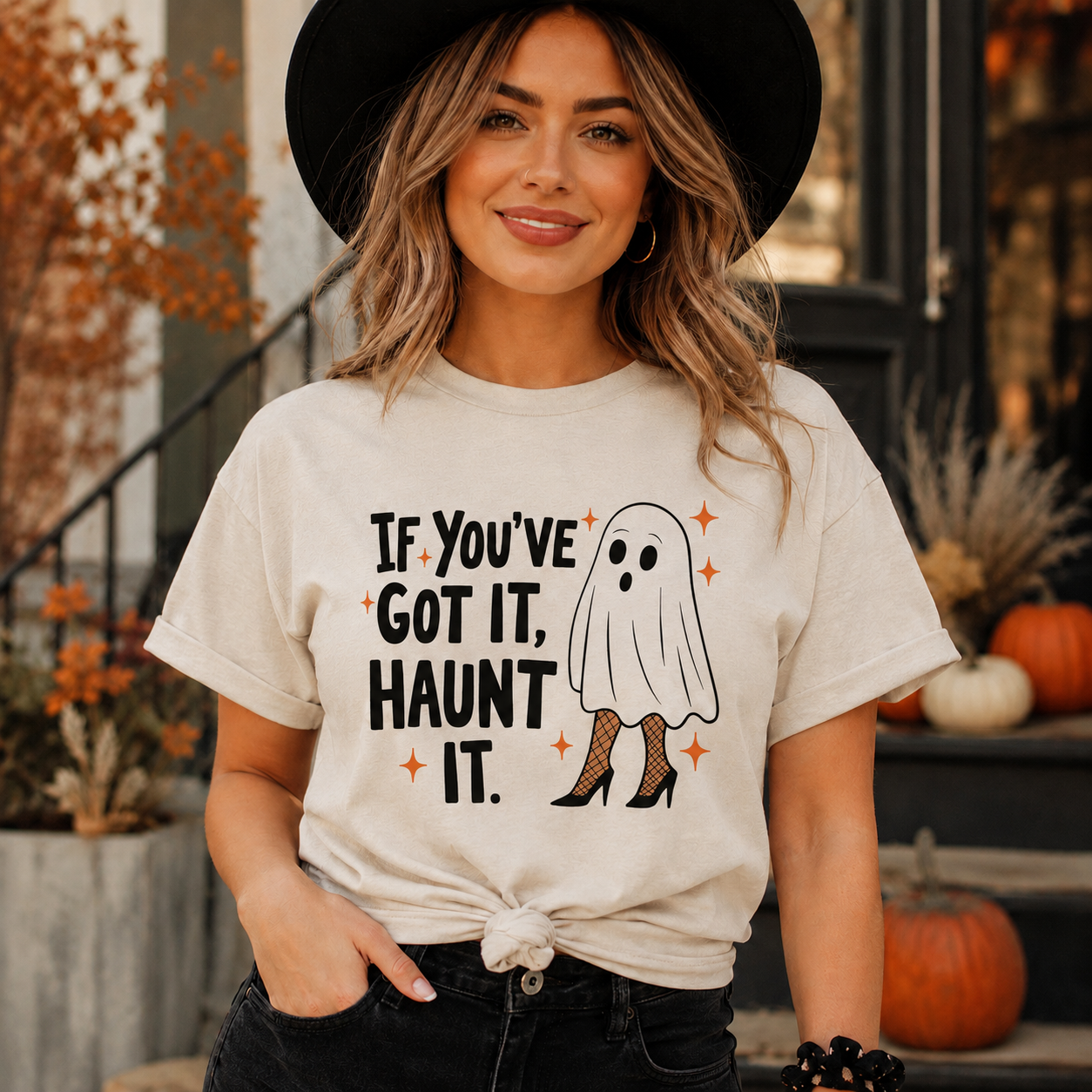 A woman wearing a cream-colored graphic t-shirt with a Halloween ghost design and the text 'IF YOU'VE GOT IT, HAUNT IT.' She is also wearing a black wide-brim hat and dark jeans, posed in front of a fall-themed storefront background.