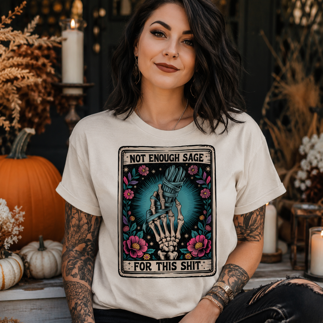 A woman wearing a cream-colored graphic t-shirt with a skeleton hand design and the text 'NOT ENOUGH SAGE FOR THIS SHIT'. She is posed in a cozy, autumn-themed setting with pumpkins, candles, and dried flowers.