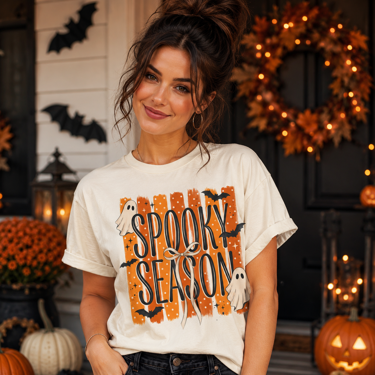 A woman wearing a white graphic t-shirt with a 'Spooky Season' Halloween design, posed in front of a decorated autumn porch with pumpkins and a wreath.