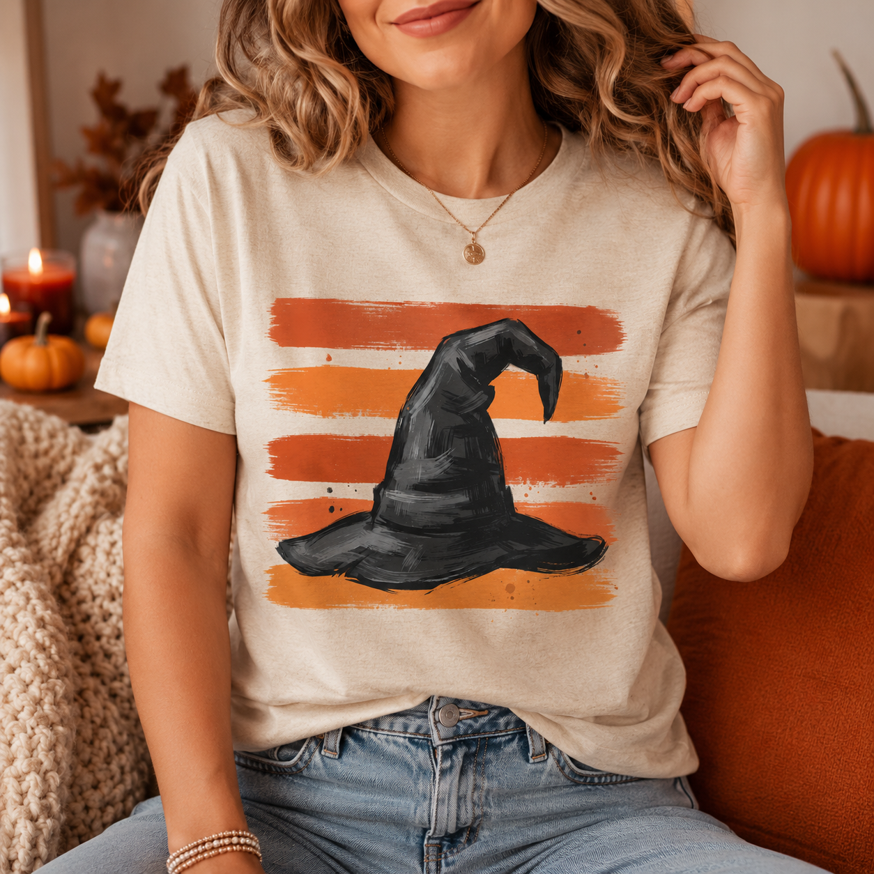 A woman wearing a beige graphic t-shirt with a black witch hat design over orange striped brushstrokes, sitting in a cozy indoor setting with pumpkins and a candle in the background.