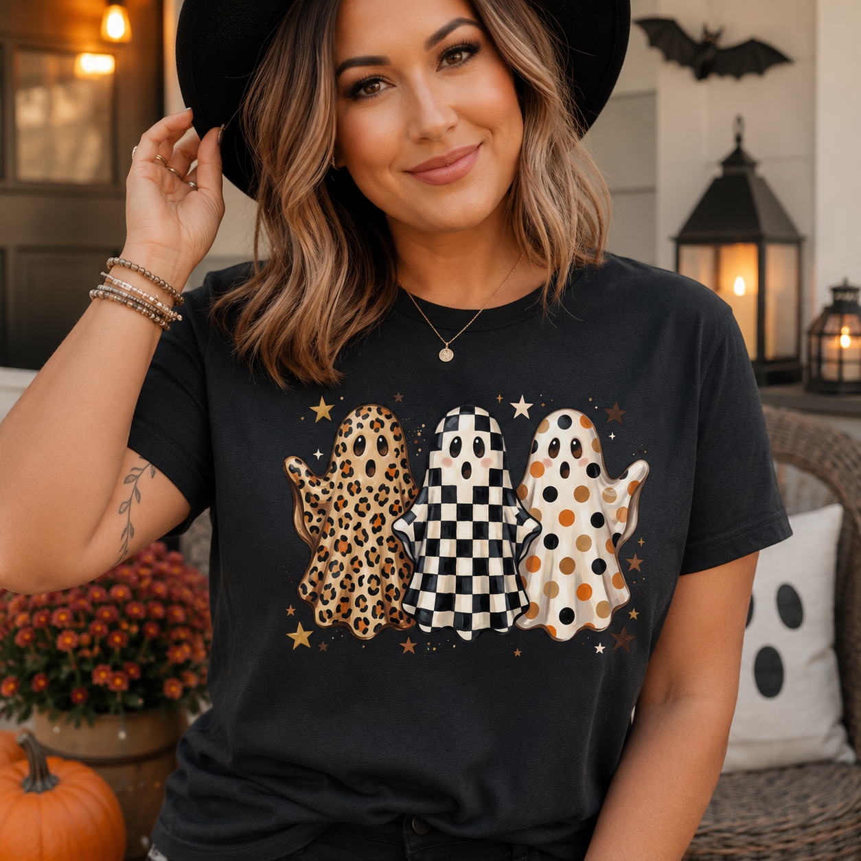 A woman wearing a black graphic t-shirt with three cartoon ghosts in leopard print, checkered pattern, and polka dot pattern, posed in a cozy outdoor patio setting.
