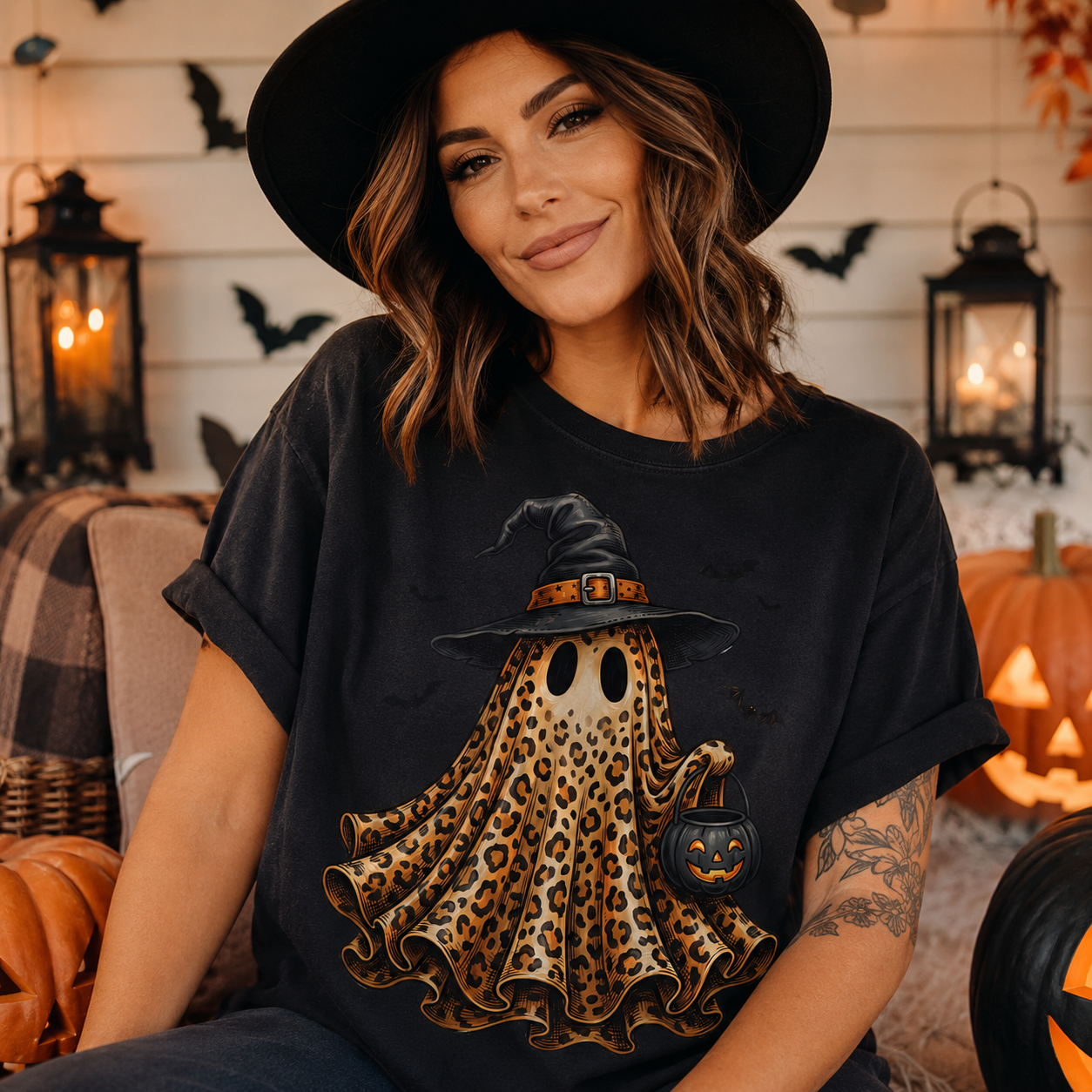 A woman wearing a black witch hat and a black graphic t-shirt featuring a leopard-print ghost with a witch hat and a pumpkin lantern, posed in a cozy Halloween-themed setting with pumpkins and lanterns.