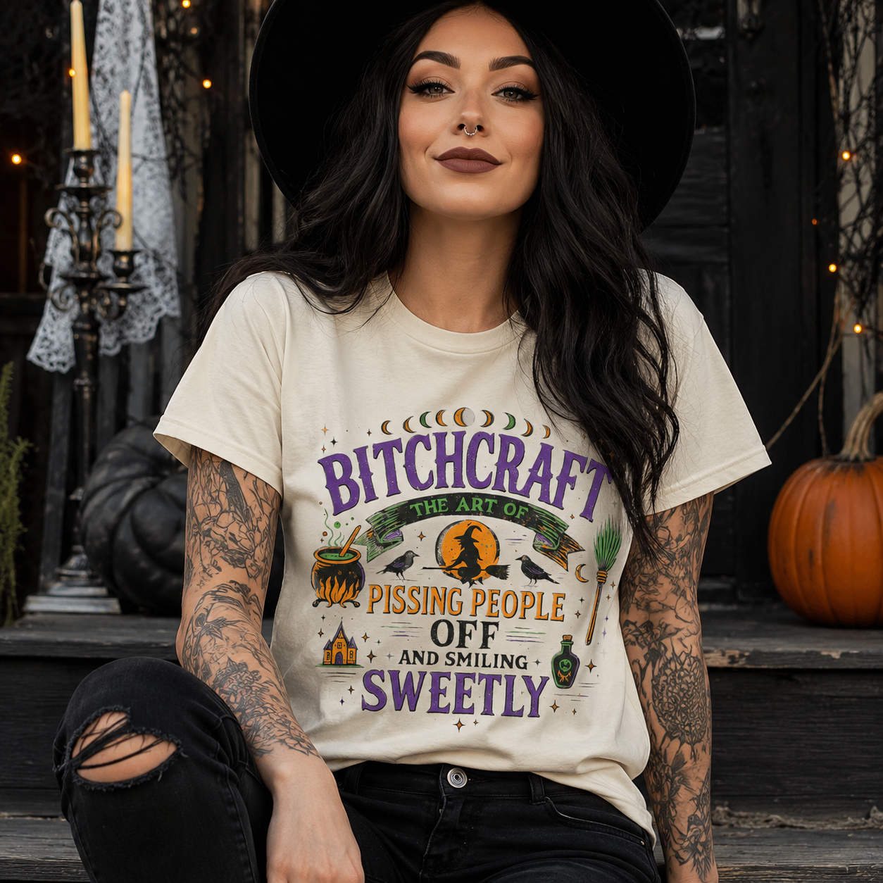 A woman wearing a cream-colored graphic t-shirt with a Halloween-themed design that reads 'Bitchcraft the Art of Pissing People Off and Smiling Sweetly'. She is wearing a black wide-brim hat and ripped black jeans, sitting on wooden steps decorated with a pumpkin and candles.