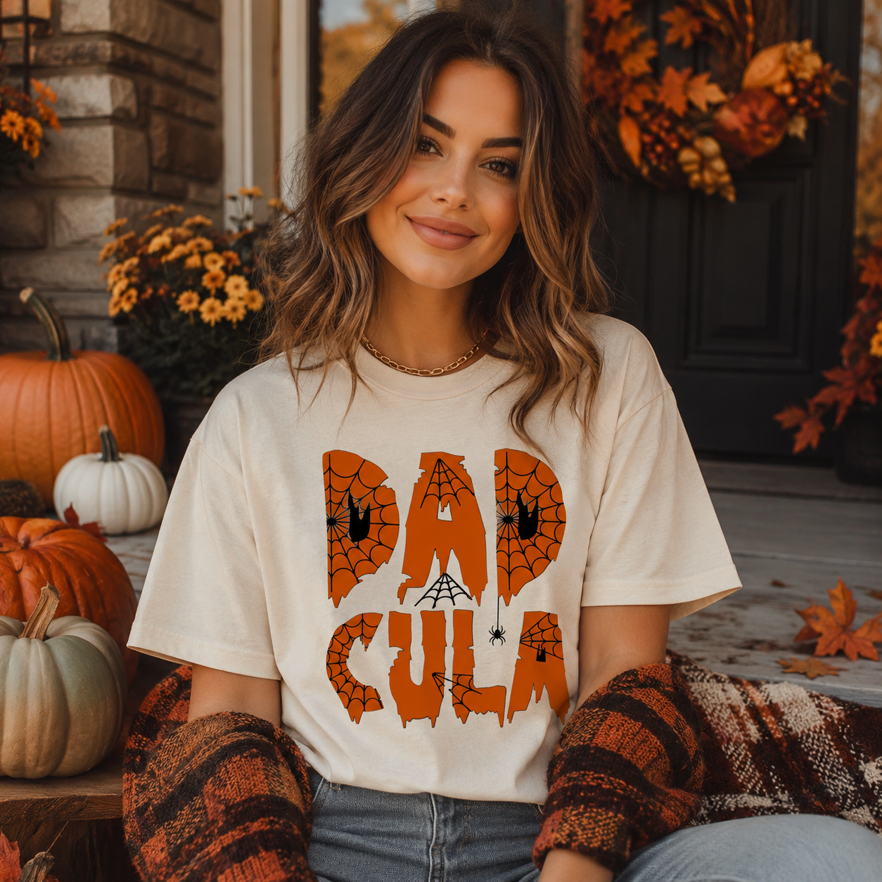 A woman wearing a cream-colored graphic t-shirt with 'DAD CULA' in orange spiderweb letters, posed on a fall-themed porch with pumpkins and autumn decorations.