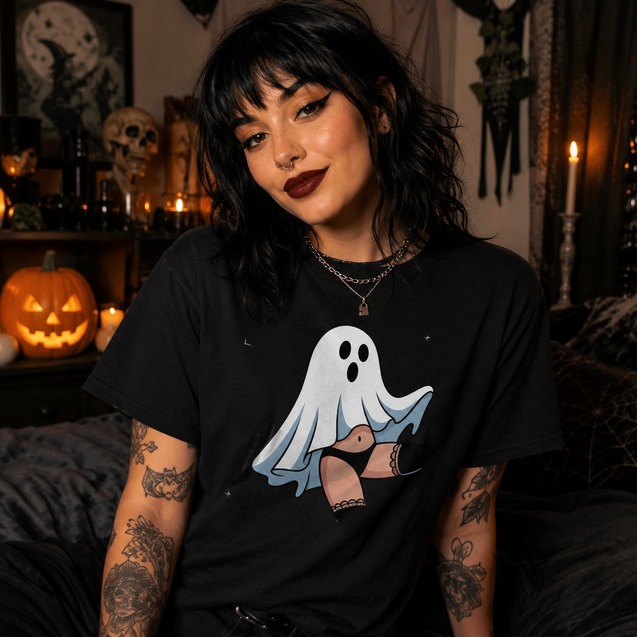 A woman with dark hair and tattoos wearing a black graphic t-shirt with a cartoon ghost illustration, posed in a cozy, dimly lit Halloween-themed room with candles, a jack-o'-lantern, and spooky decorations.