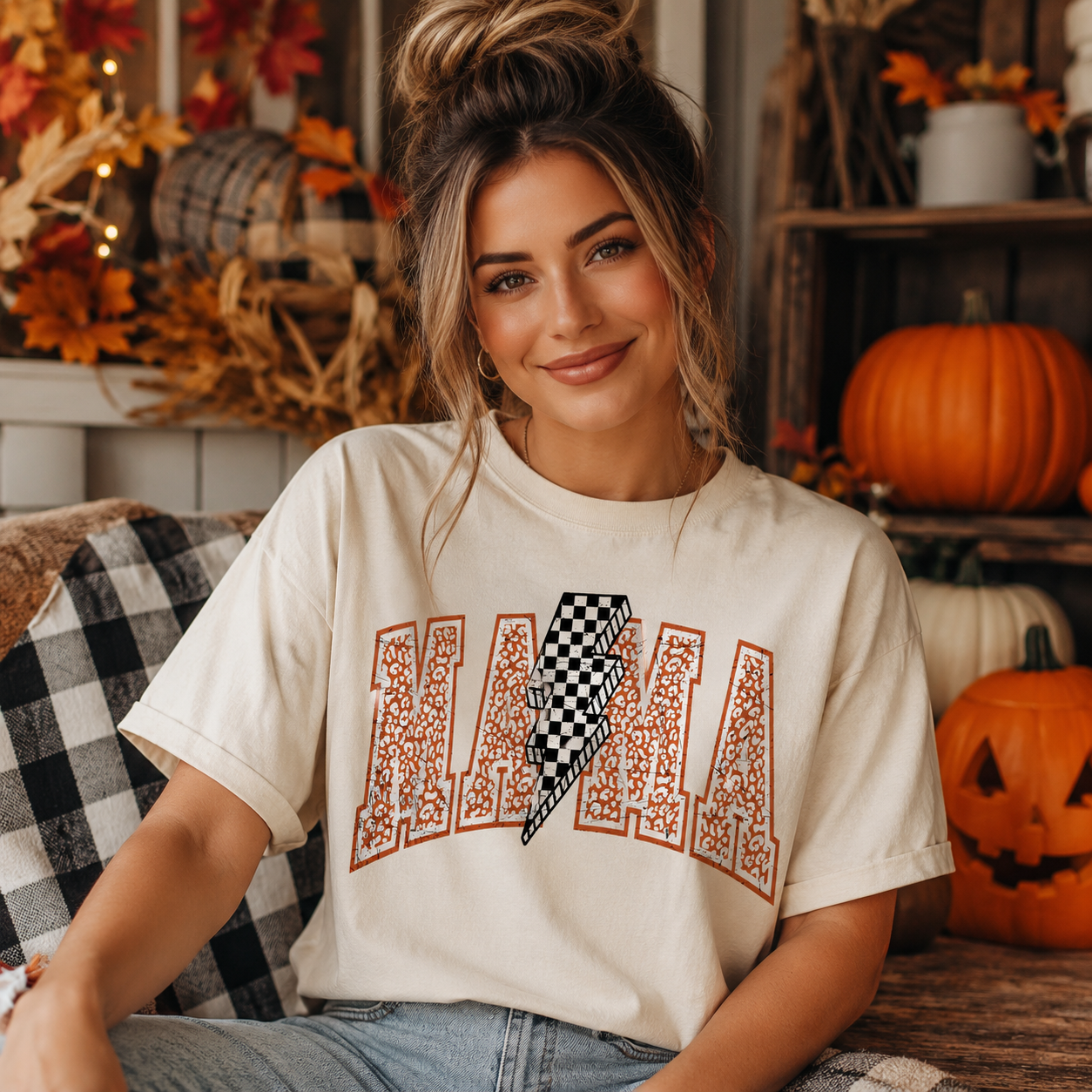 A woman wearing a cream-colored graphic t-shirt with a brown arch design spelling 'MAMA' and a black-and-white lightning bolt in the center, posed in a cozy autumn-themed room with pumpkins and fall decorations.