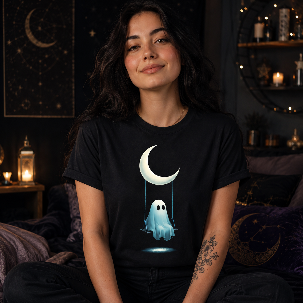 A woman wearing a black t-shirt with a cute graphic of a white crescent moon and a glowing blue ghost, sitting in a cozy, dark-themed bedroom with candles and celestial wall art.