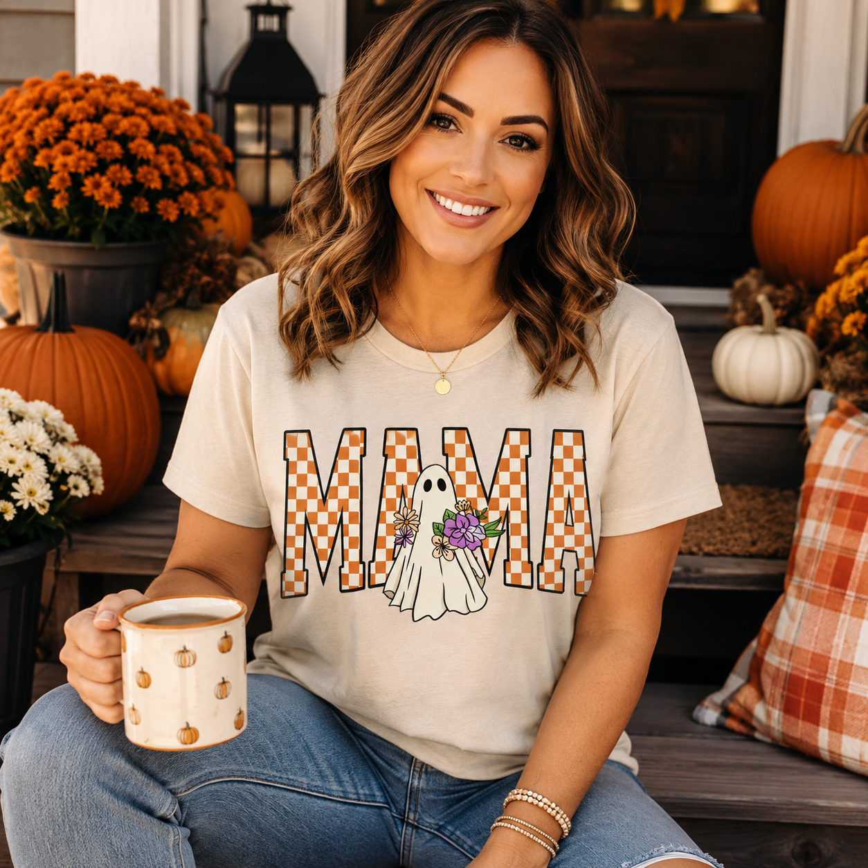 A woman sitting on a porch steps wearing a beige graphic t-shirt with 'MAMA' in orange and white checkered letters, featuring a white ghost holding a purple flower. She is holding a matching pumpkin-patterned mug.