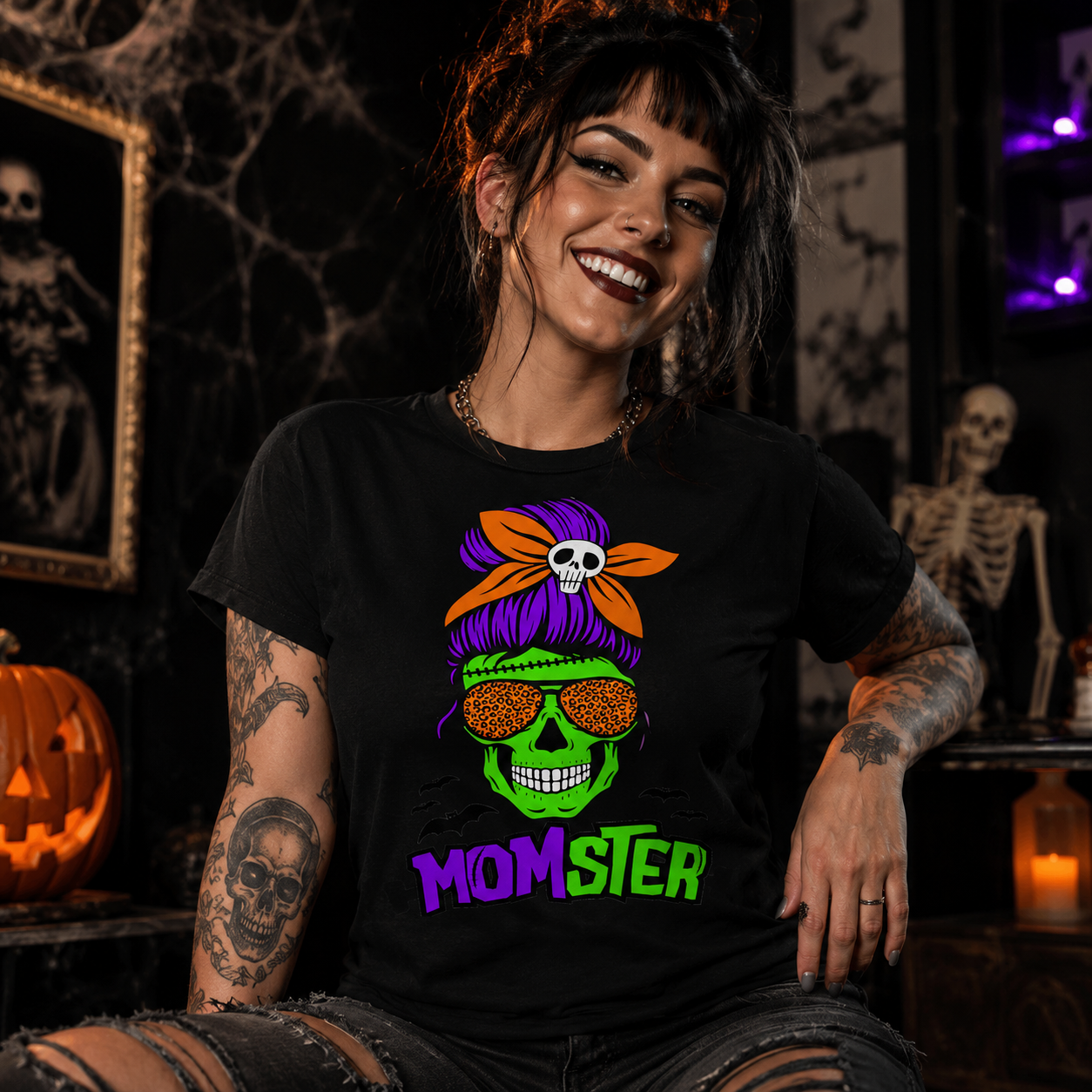 A woman wearing a black graphic t-shirt with a colorful 'MOMSTER' Halloween design, posed in a spooky, dark room with skeletons and a pumpkin.