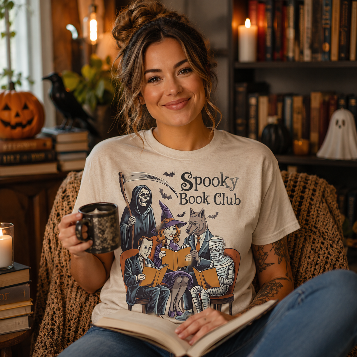 A woman sitting in a cozy room wearing a cream-colored graphic t-shirt that reads 'Spooky Book Club' with a Halloween-themed illustration of a grim reaper, witch, cat, and mummy reading together. She is holding a mug and an open book.