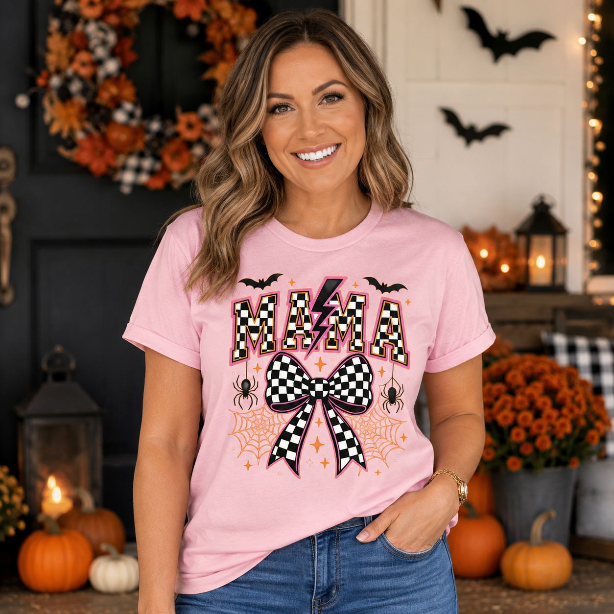 A woman wearing a light pink graphic t-shirt with a Halloween-themed 'MAMA' design featuring a checkered bow, spiders, bats, and pumpkins in the background.