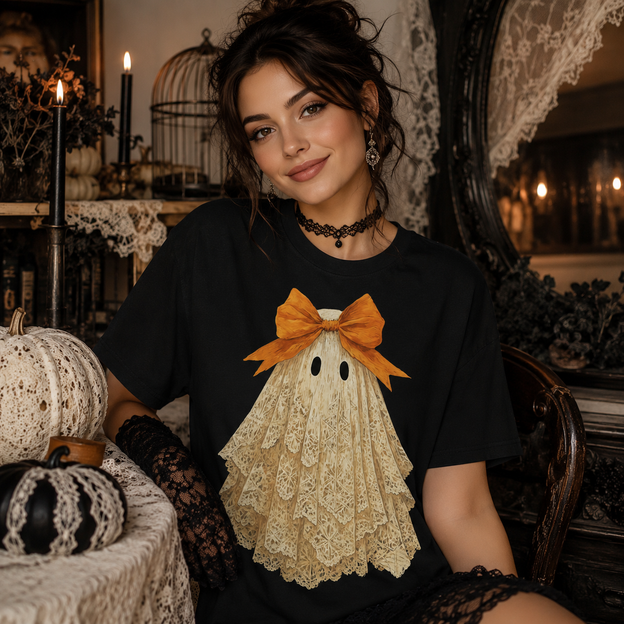 A woman wearing a black t-shirt with a lace-embroidered ghost graphic and an orange bow, posed in a cozy, dark, vintage-style room with pumpkins and candles.