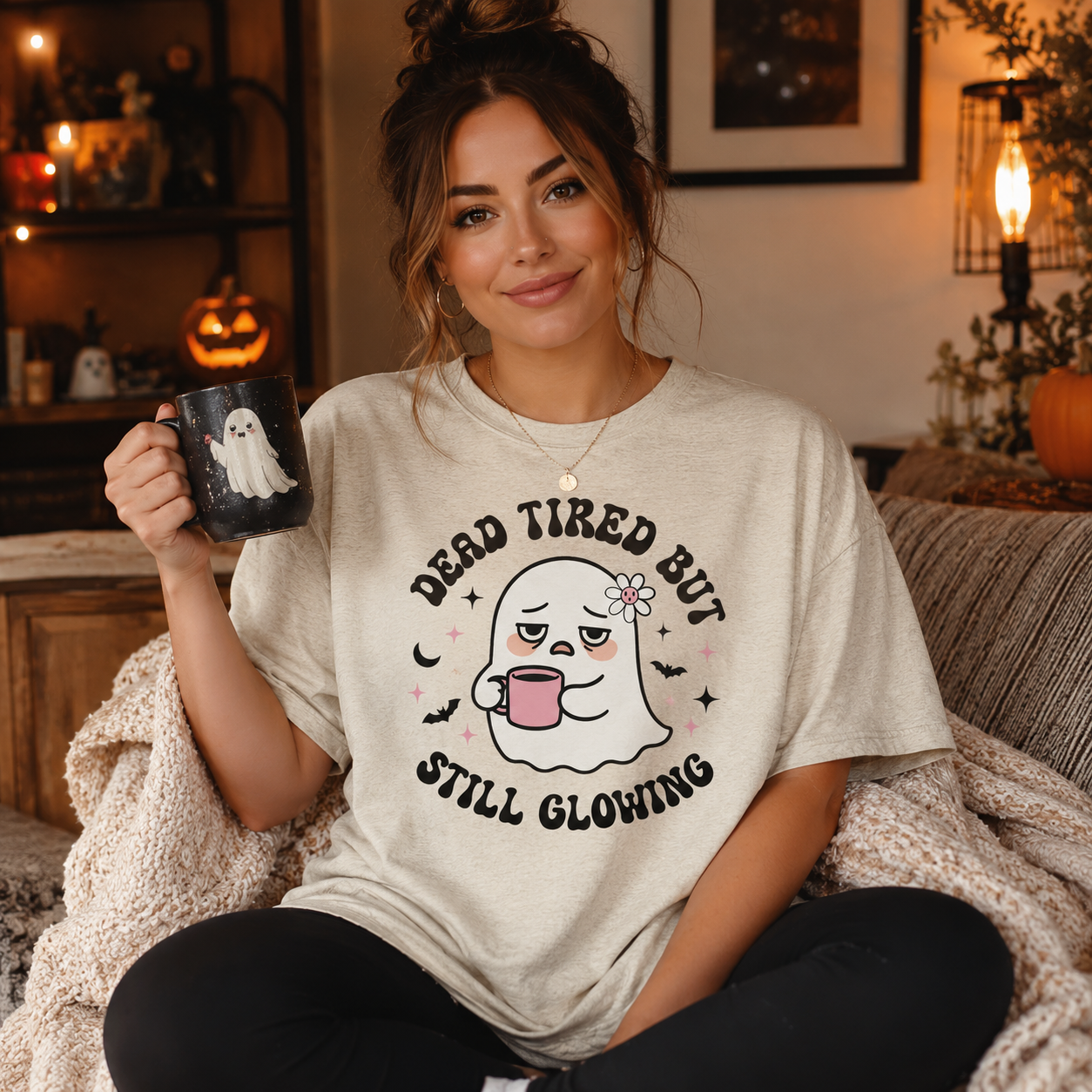 A woman sitting on a couch in a cozy living room setting, wearing a light beige graphic t-shirt with a cartoon ghost design and the text 'DEAD TIRED BUT STILL GLOWING'. She is holding a black mug with a ghost graphic.