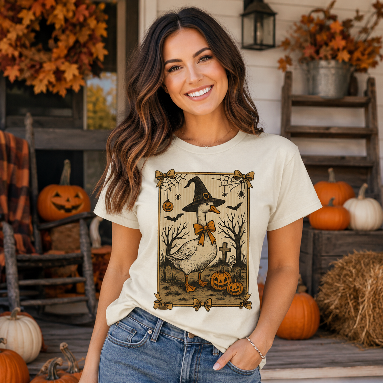 A woman stands on a rustic fall-themed porch wearing a cream-colored graphic t-shirt with a Halloween goose design, paired with blue jeans. The background features pumpkins, hay, and autumn decorations.