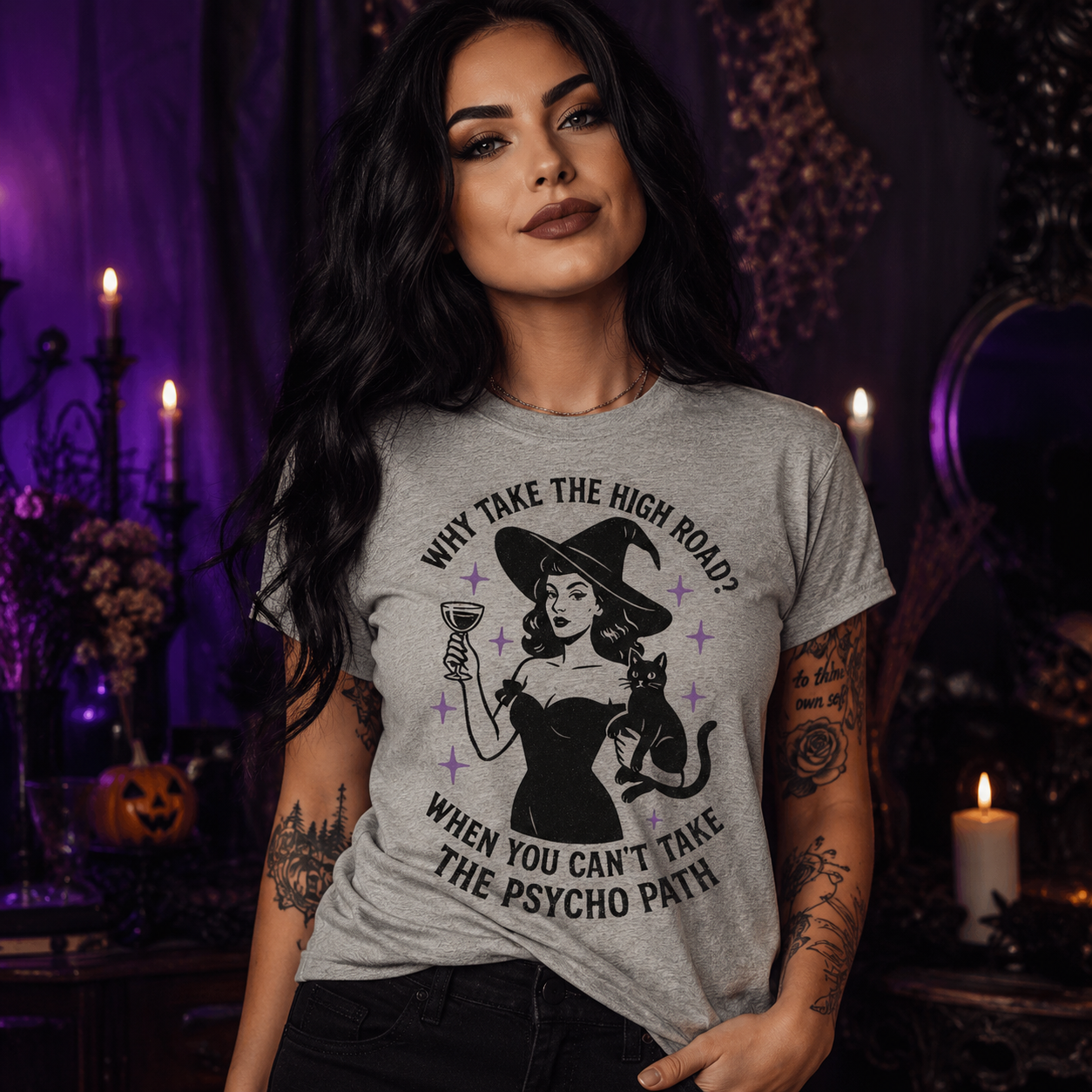 A woman wearing a light gray graphic t-shirt with a black witch-themed print that reads 'WHY TAKE THE HIGH ROAD? WHEN YOU CAN'T TAKE THE PSYCHO PATH!' in a dark, atmospheric Halloween setting with candles and pumpkins.