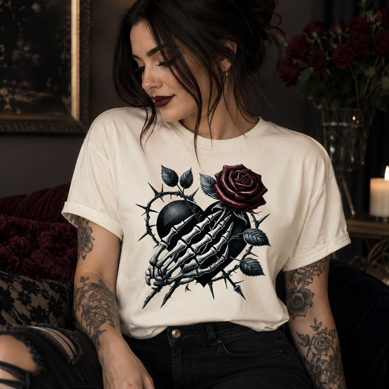 A woman wearing a cream-colored graphic t-shirt with a black skeletal hand and a dark red rose design, posed in a dimly lit, moody room.
