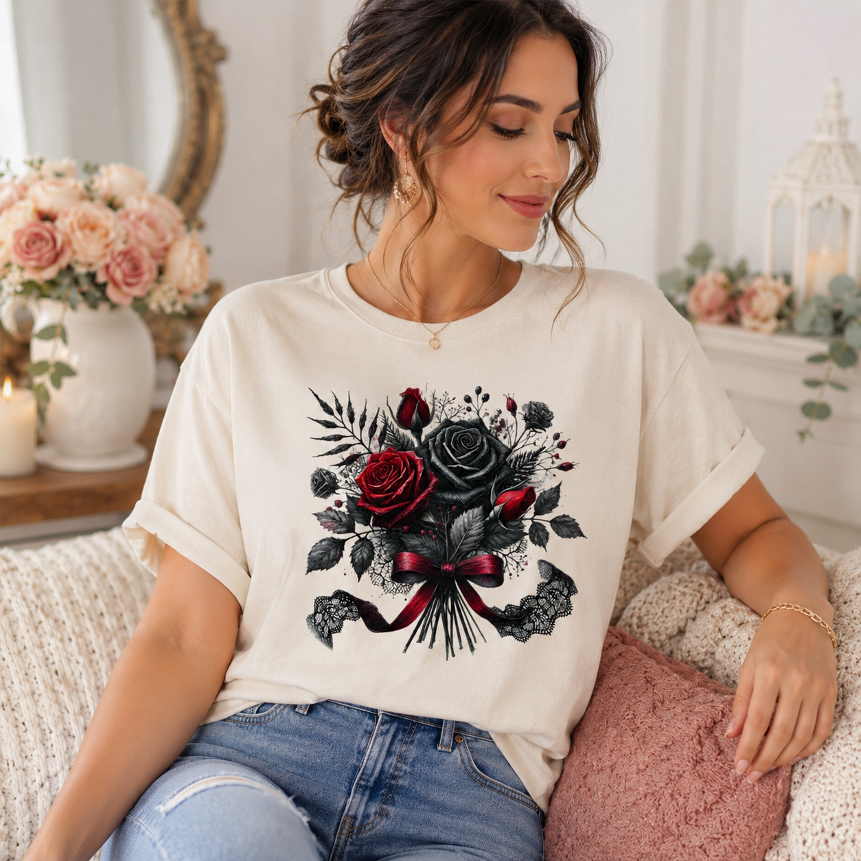 A woman wearing a cream-colored t-shirt with a gothic-style floral graphic of black and red roses, lace, and a ribbon bow. She is sitting on a cozy couch in a beautifully decorated room with pink flowers and candles.