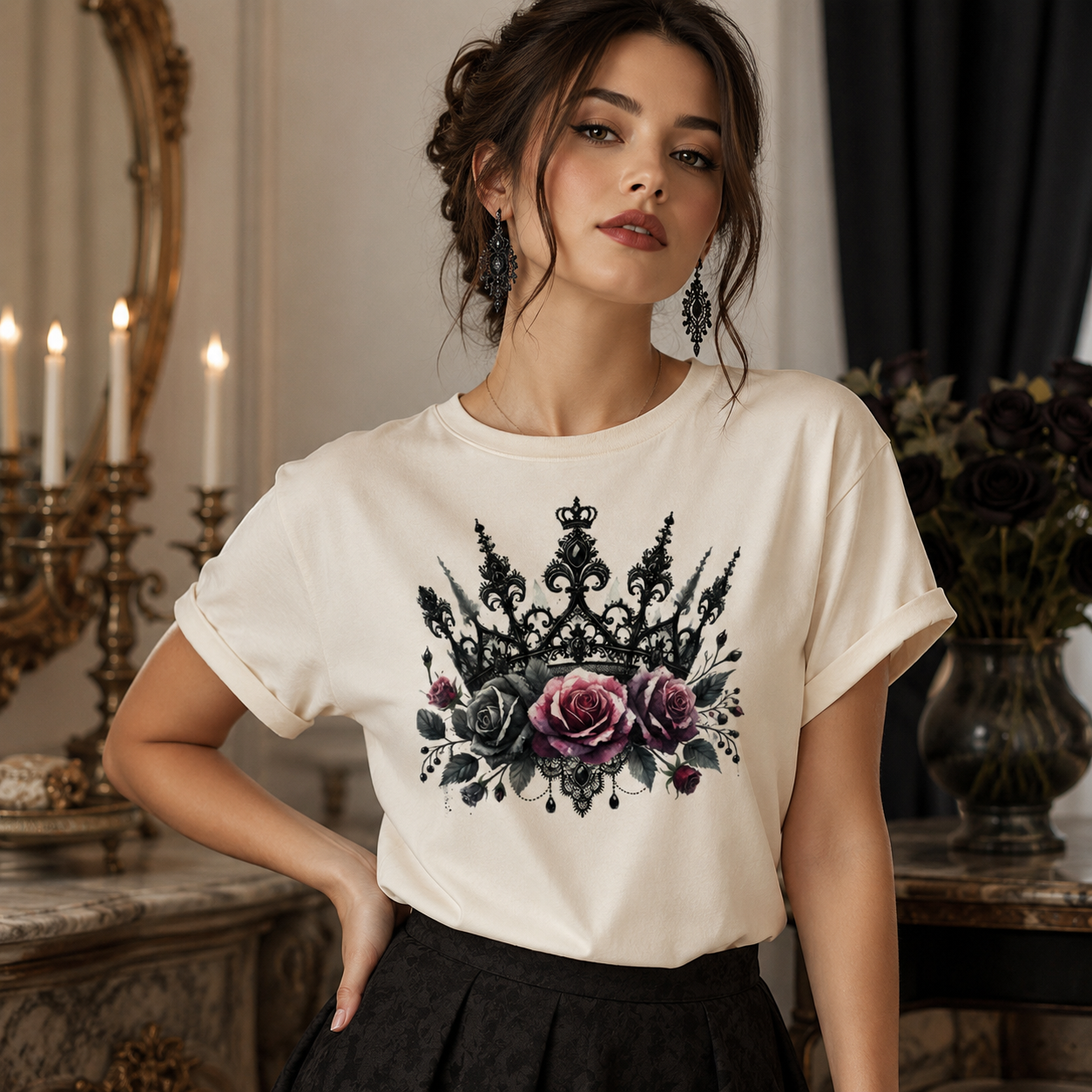 A woman wearing a cream-colored t-shirt with a black crown and purple rose graphic print, paired with a black skirt, posed in an elegant indoor setting with candles and a mirror in the background.