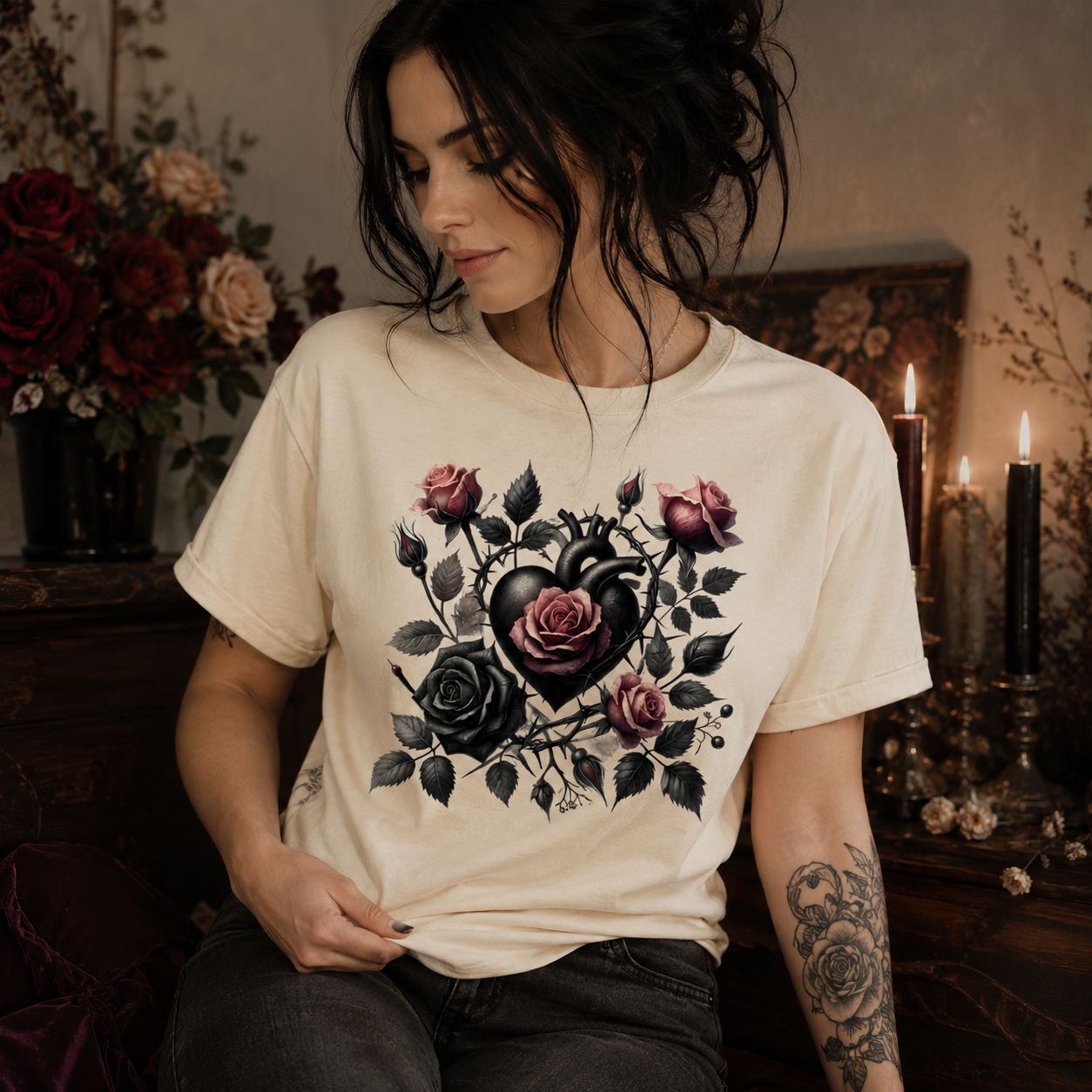 A woman wearing a cream-colored graphic t-shirt with a black and pink rose heart design, posed in a moody, dimly lit room with candles and flowers.