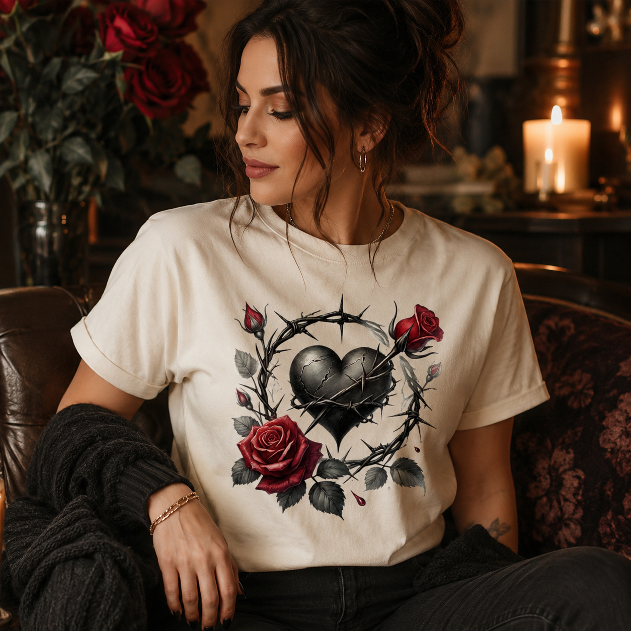 A woman wearing a cream-colored graphic t-shirt with a black heart, thorny wreath, and red roses design, sitting in a cozy, dimly lit room with candles and flowers in the background.