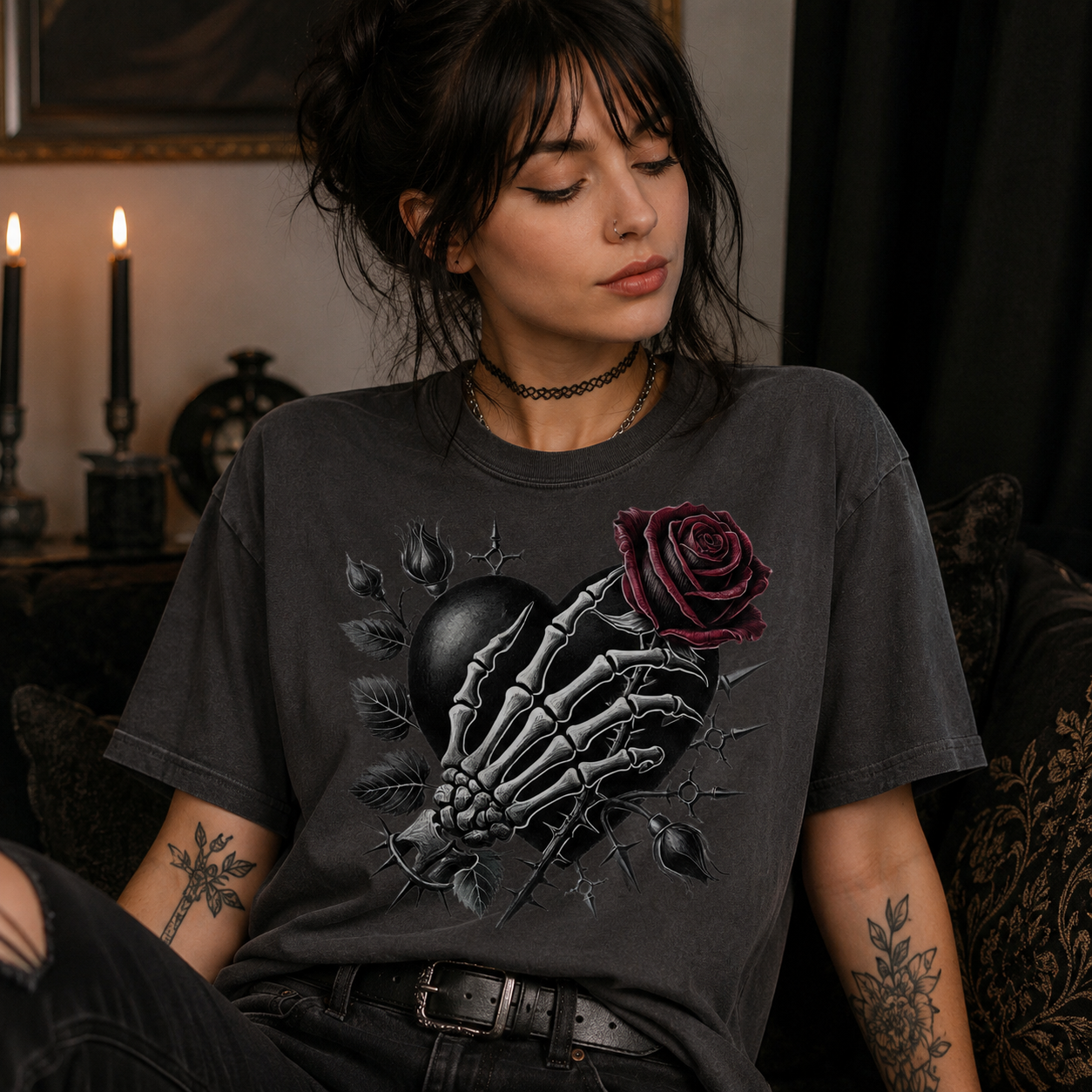 A woman wearing a dark charcoal gray graphic t-shirt with a skeleton hand holding a dark red rose, seated in a dimly lit, moody room with candles.