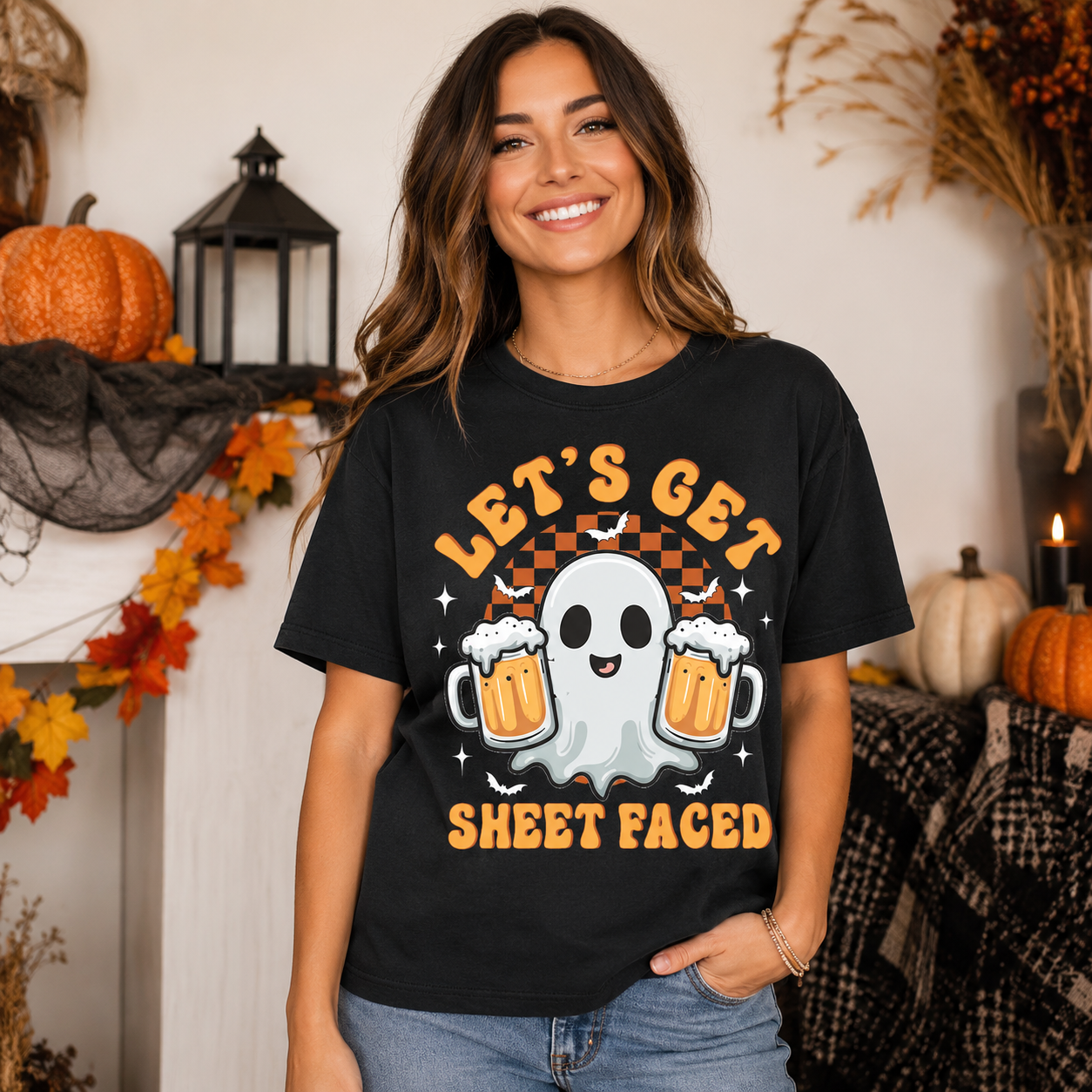 A woman wearing a black graphic t-shirt with a Halloween design that reads 'LET'S GET SHEET FACED' in orange text, featuring a white ghost and two beer mugs. The background is a cozy, decorated autumn scene with pumpkins, a lantern, and fall foliage.
