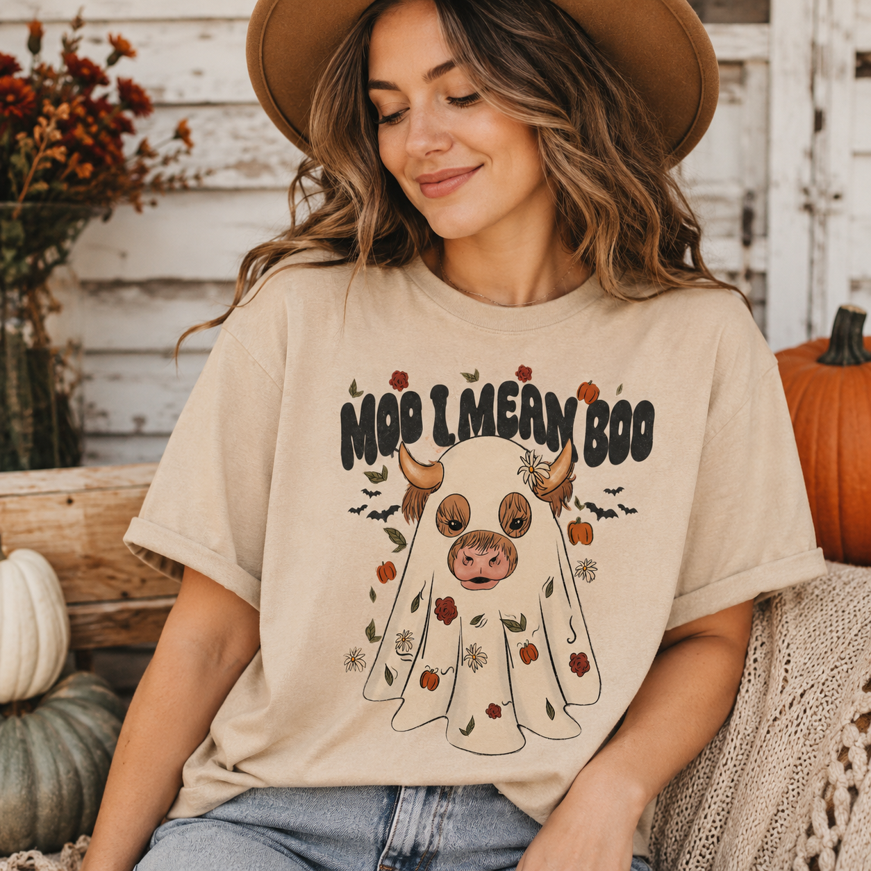 A woman wearing a beige graphic t-shirt with a cartoon ghost and the text 'Moo Lmeran Boo' in a rustic, autumn-themed setting with pumpkins and a wooden fence.